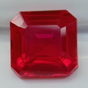 Puede incluir: Una gema roja oscura tallada en forma rectangular. La superficie pulida de la gema tiene múltiples facetas que reflejan la luz, creando un brillo brillante. La piedra preciosa es de un color rojo vibrante y saturado.