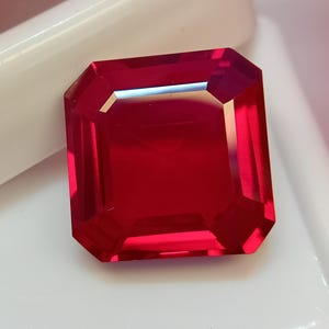 May include: A faceted, rectangular-cut, deep red gemstone. The gem has a polished surface with sharp, defined edges. The gem is set against a white background.