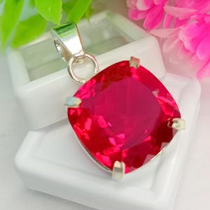 May include: A square-cut, vibrant red gemstone pendant set in a silver-toned metal prong setting. The pendant features a faceted surface that reflects light, attached to a polished silver-toned bail at the top for hanging on a chain or cord.