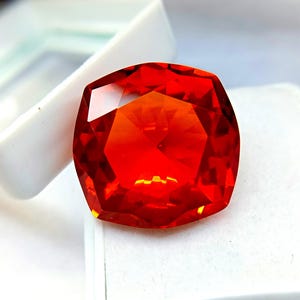May include: A faceted, cushion-cut gemstone with a vibrant orange-red hue. The gem is set against a white background, showcasing its clarity and brilliance. The gemstone's facets reflect light, creating a dazzling display.