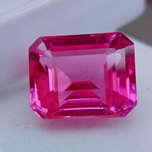 Puede incluir: Una vibrante gema rosa de corte rectangular. La gema tiene una superficie facetada que refleja la luz, mostrando su claridad y brillo. El color es un rosa intenso y saturado, con un acabado pulido. La gema se encuentra sobre un fondo blanco.