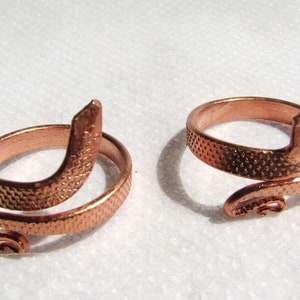 May include: Two copper rings shaped like snakes with textured surfaces. The rings are coiled and have a tail that curls up.