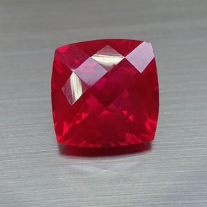 Puede incluir: Una gema facetada, de corte cuadrado, en un tono rojo intenso y vibrante. La superficie de la gema refleja la luz, mostrando sus múltiples facetas y su brillo interno. La gema se encuentra sobre un fondo metálico neutro.