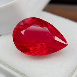 May include: A vivid red, pear-cut gemstone. The gem is displayed against a white backdrop, highlighting its brilliant facets and rich colour. The stone's cut reflects light, producing a sparkling effect. The gem is likely a ruby.