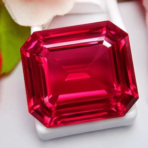 May include: A vibrant, deep red gemstone cut in a rectangular emerald shape with sharp, clean facets. The stone displays high clarity and intense saturation, reflecting light across its polished surface while resting on a white base.
