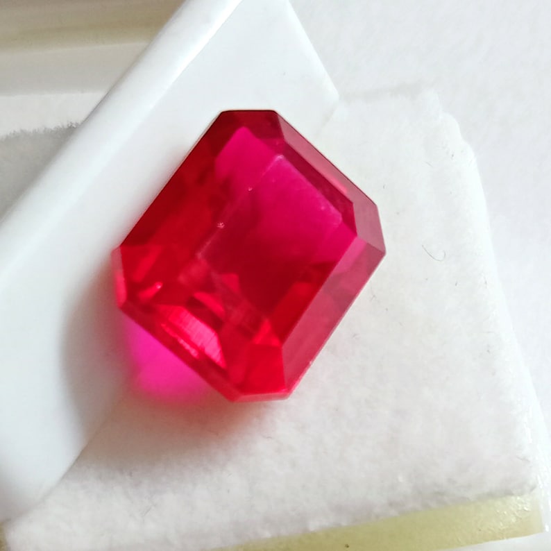 May include: A vibrant, deep red gemstone with a rectangular emerald cut. The gemstone is faceted and has a high polish.