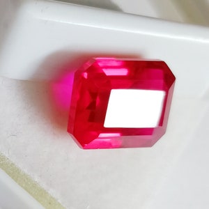 May include: A vibrant, deep red gemstone with a rectangular emerald cut. The gemstone is faceted and has a high polish.