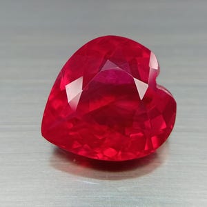 Puede incluir: Una gema facetada en forma de corazón, de un tono rojo vibrante. La superficie de la gema refleja la luz, mostrando sus múltiples facetas y estructura interna. La gema pulida se encuentra sobre un fondo neutro.