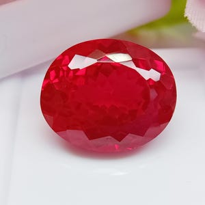 Puede incluir: Una gema roja intensa, de forma ovalada y tallada en facetas. La gema tiene un corte brillante, que refleja la luz en sus múltiples facetas. La piedra se encuentra sobre un fondo blanco, con suaves acentos rosas y verdes en el fondo.