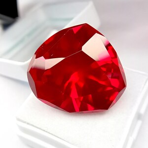 May include: A large, faceted, heart-shaped, deep red gemstone. The gem is displayed on a white surface inside a white box, with the facets reflecting light. The gemstone's colour is a vibrant, saturated red.