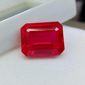 Peut inclure: Une pierre précieuse rubis rectangulaire et facettée, d'un rouge vif. La gemme est posée sur un fond blanc, mettant en valeur sa clarté et sa brillance. La taille de la pierre met en évidence sa structure interne.