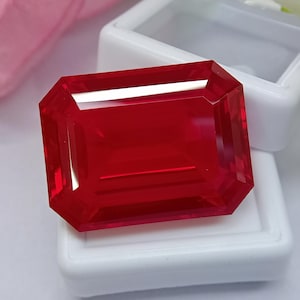 Könnte beinhalten: Ein leuchtend roter, transparenter Edelstein im rechteckigen Smaragdschliff mit facettierten Kanten. Der Stein zeigt einen tiefen karmesinroten Farbton und hohe Klarheit, wobei er das Licht auf seinen polierten Oberflächen reflektiert.