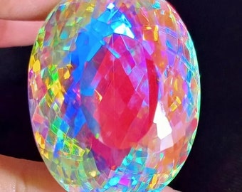 May include: A large, oval-shaped, iridescent gemstone with a faceted cut. The gemstone is clear with a rainbow of colors, including red, blue, green, and yellow. The gemstone is held in a hand.