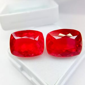 Op de afbeelding: Twee gefacetteerde, kussenvormig geslepen, helderrode edelstenen. De edelstenen worden getoond op een wit oppervlak, met een witte container op de achtergrond. De edelstenen hebben een hoge mate van helderheid en glans.