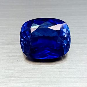 Peut inclure: Une pierre pr&eacute;cieuse saphir taill&eacute;e en coussin. Le saphir est d'un bleu profond et vibrant, avec de multiples facettes qui r&eacute;fl&eacute;chissent la lumi&egrave;re. La pierre est pos&eacute;e sur un fond neutre.