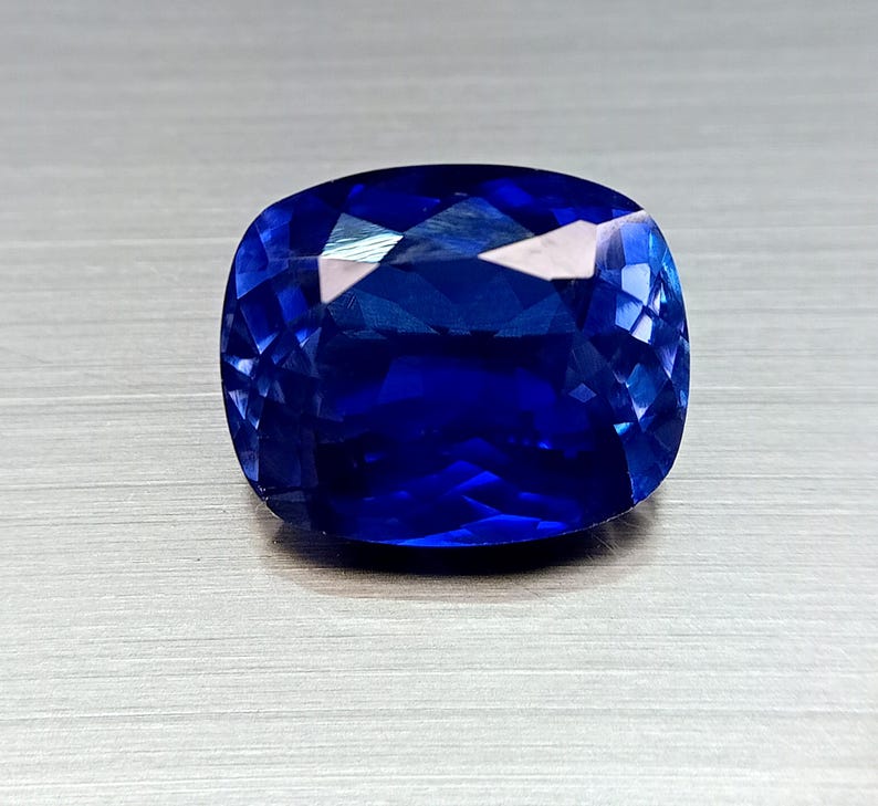 Peut inclure: Une pierre pr&eacute;cieuse saphir taill&eacute;e en coussin. Le saphir est d'un bleu profond et vibrant, avec de multiples facettes qui r&eacute;fl&eacute;chissent la lumi&egrave;re. La pierre est pos&eacute;e sur un fond neutre.