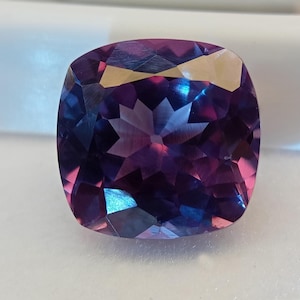 May include: A faceted, cushion-cut gemstone with a color-changing effect. The gem displays shades of purple, blue, and pink, with a star-like pattern visible in the center. The stone is set against a white background.