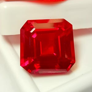 May include: A square-cut, vibrant red gemstone with faceted surfaces that reflect light. The stone features sharp corners and a transparent, deep crimson color, displaying internal clarity and a polished finish throughout its geometric structure.