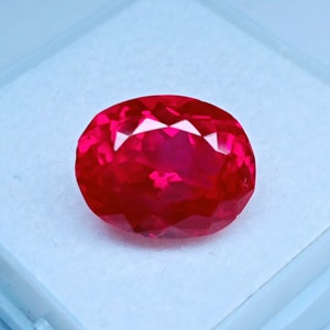 May include: A faceted, oval-shaped, vibrant red gemstone. The gem has a brilliant cut with multiple facets that reflect light. The stone is set against a white background, highlighting its color and clarity. This gemstone is suitable for jewelry.