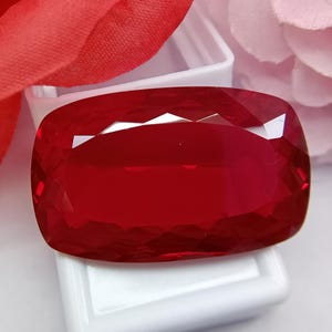 May include: A faceted, rectangular, deep red gemstone. The gem is set against a white background, with a red fabric and a pink flower in the background. The gem has many facets that reflect light.