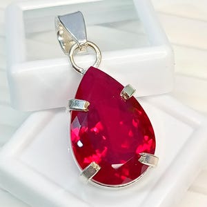 May include: A pear-shaped, faceted deep red gemstone pendant set in a silver-toned metal prong setting. The pendant features a polished silver-toned bail at the top, designed for attachment to a chain, showcasing a vibrant, translucent red hue.
