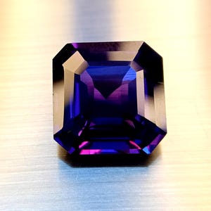May include: A faceted gemstone with a rectangular emerald cut. The gem displays a gradient of colors, transitioning from deep purple to vibrant blue and hints of pink. The polished facets reflect light, creating a sparkling effect.