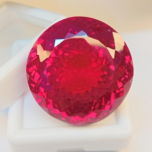 Peut inclure: Une grosse pierre précieuse ronde et facettée, d'un rouge profond. La gemme est présentée dans un boîtier en plastique blanc, mettant en valeur sa taille brillante et ses surfaces réfléchissantes. Les facettes de la gemme captent la lumière.