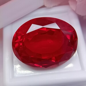 Könnte beinhalten: Ein facettierter, oval geformter, tiefroter Edelstein. Der Edelstein ist vor einem weißen Hintergrund platziert, der seinen Brillantschliff und seine lebendige Farbe zeigt. Die Facetten des Edelsteins reflektieren das Licht. Es ist ein Rubin.