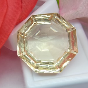 May include: A faceted, octagonal gemstone with a pale yellow hue. The gem is set against a white surface, with a red fabric and a pink fabric in the background. The gemstone's facets reflect light, creating a sparkling effect.