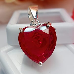 May include: A heart-shaped pendant with a large, faceted, deep red gemstone. The gemstone is held in place by silver-colored prongs and a silver bail. The pendant is displayed against a white background.