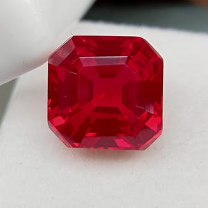 May include: A faceted, square-cut, bright red gemstone. The gem has a symmetrical octagonal shape with multiple facets that reflect light. The gemstone is set against a white background.