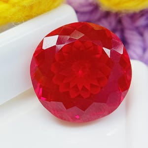 May include: A round, faceted gemstone in a vibrant, translucent red colour. The stone features a complex, symmetrical geometric cut on its surface, creating a star-like pattern that reflects light across its polished, multi-faceted circular face.