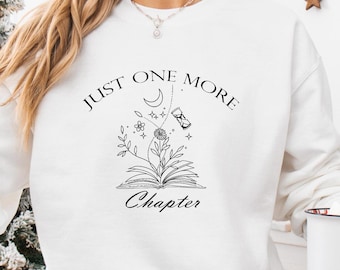 May include: White sweatshirt with a black graphic design that says "Just One More Chapter" with a crescent moon, hourglass, and stylized flowers above an open book.