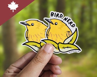 Può includere: Un adesivo con due uccelli gialli dei cartoni animati con la scritta "BIRD NERD". Gli uccelli sono posati su foglie verdi. L'adesivo è tenuto in mano, con la scritta "Made in Canada!" e un logo a forma di foglia d'acero.