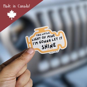 May include: A sticker in the shape of a car's engine light, with the text "THIS LITTLE LIGHT OF MINE I'M GONNA LET IT SHINE" in black. The sticker is yellow and white, with a red and white "Made in Canada!" logo in the upper left corner.