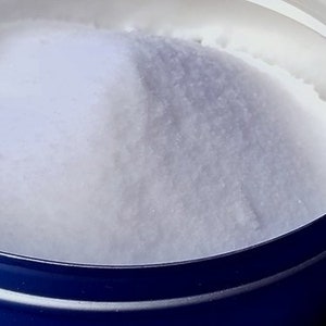 May include: Close-up of a white, granular substance filling a blue and silver container. The substance appears to be a fine powder, possibly salt or sugar, with a slightly uneven surface. The container's rim is visible.