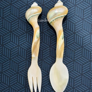 May include: A set of two shell spoons, one with a fork and one with a spoon head. The spoons are made from natural shell and have a brown and white swirl pattern. The text "SilkGardenArt" is visible on the spoons.