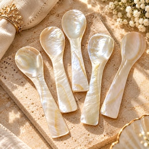 May include: A set of five small spoons crafted from iridescent mother-of-pearl, each featuring a smooth, polished surface with natural cream and white swirling patterns. The spoons have a rounded bowl and a flat, tapered handle, showcasing the unique luster.