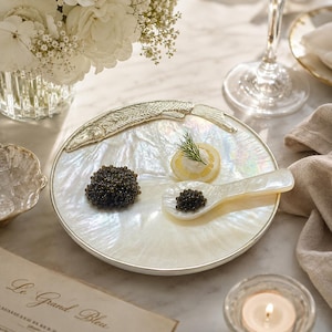 May include: A round serving plate made of iridescent mother-of-pearl with a silver-toned metal fish ornament along the top edge. The plate holds a small mound of black caviar, a lemon slice topped with cream and dill, and a mother-of-pearl spoon.