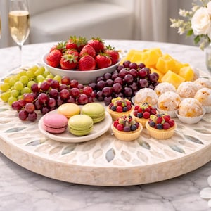May include: A circular serving board with a mosaic pattern, displaying a selection of fresh fruit, pastries, and macarons. Strawberries, grapes, and pineapple are arranged with mini fruit tarts and sugared pastries. Two champagne flutes are visible.