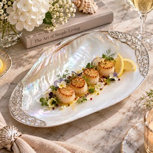 May include: An oval serving platter made of iridescent mother-of-pearl with ornate silver-toned metal accents on the edges. The platter holds a dish of seared scallops garnished with fresh herbs, edible flowers, and thin lemon slices.