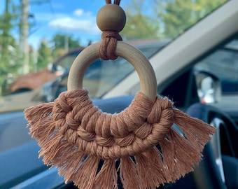 May include: A brown and beige macrame car charm with a wooden ring and bead. The charm is hanging from a rearview mirror in a car.