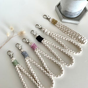 Könnte beinhalten: Fünf weiße Makramee-Schlüsselanhänger mit verschiedenen farbigen Akzenten. Die Schlüsselanhänger sind aus einem dicken, weißen Seil gefertigt und haben einen silbernen Metallverschluss. Die Akzente sind in den Farben Grün, Rosa, Grau, Schwarz und Gelb.