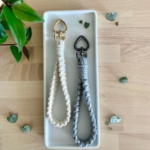 May include: Two handmade macrame keychains with heart-shaped metal clasps. One keychain is white and the other is gray. The keychains are displayed on a white rectangular plate.