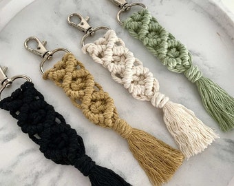 May include: Four macrame keychains with tassels in black, beige, mustard yellow, and sage green. The keychains are hanging on a white marble surface.