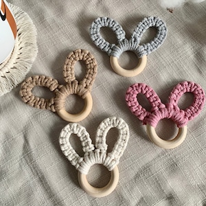 May include: Four crocheted bunny-shaped teethers with wooden rings. The teethers are in grey, brown, white, and pink.