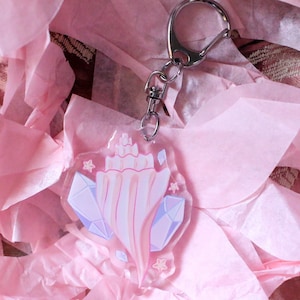 May include: A pink and white seashell charm with blue crystals and silver stars on a clear acrylic keychain with a silver metal ring.