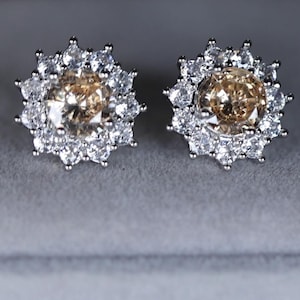 May include: A pair of stud earrings featuring a central, round, light brown gemstone surrounded by a halo of smaller, clear gemstones. The earrings are set in a silver-colored metal and are displayed on a gray surface.