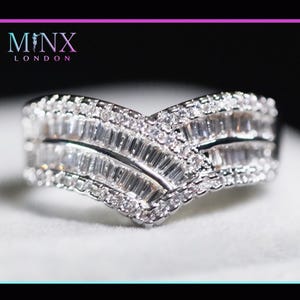 Peut inclure: Une bague argentée en forme de V, incrustée de nombreux petits pierres précieuses transparentes. La bague présente une construction en couches avec un mélange de pierres de taille baguette et rondes, créant un effet scintillant. La marque "MINX LONDON" est visible.
