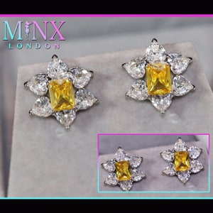 Puede incluir: Un par de pendientes de color amarillo y blanco con forma de flor con una piedra amarilla cuadrada en el centro. Los pendientes están sobre un fondo blanco.